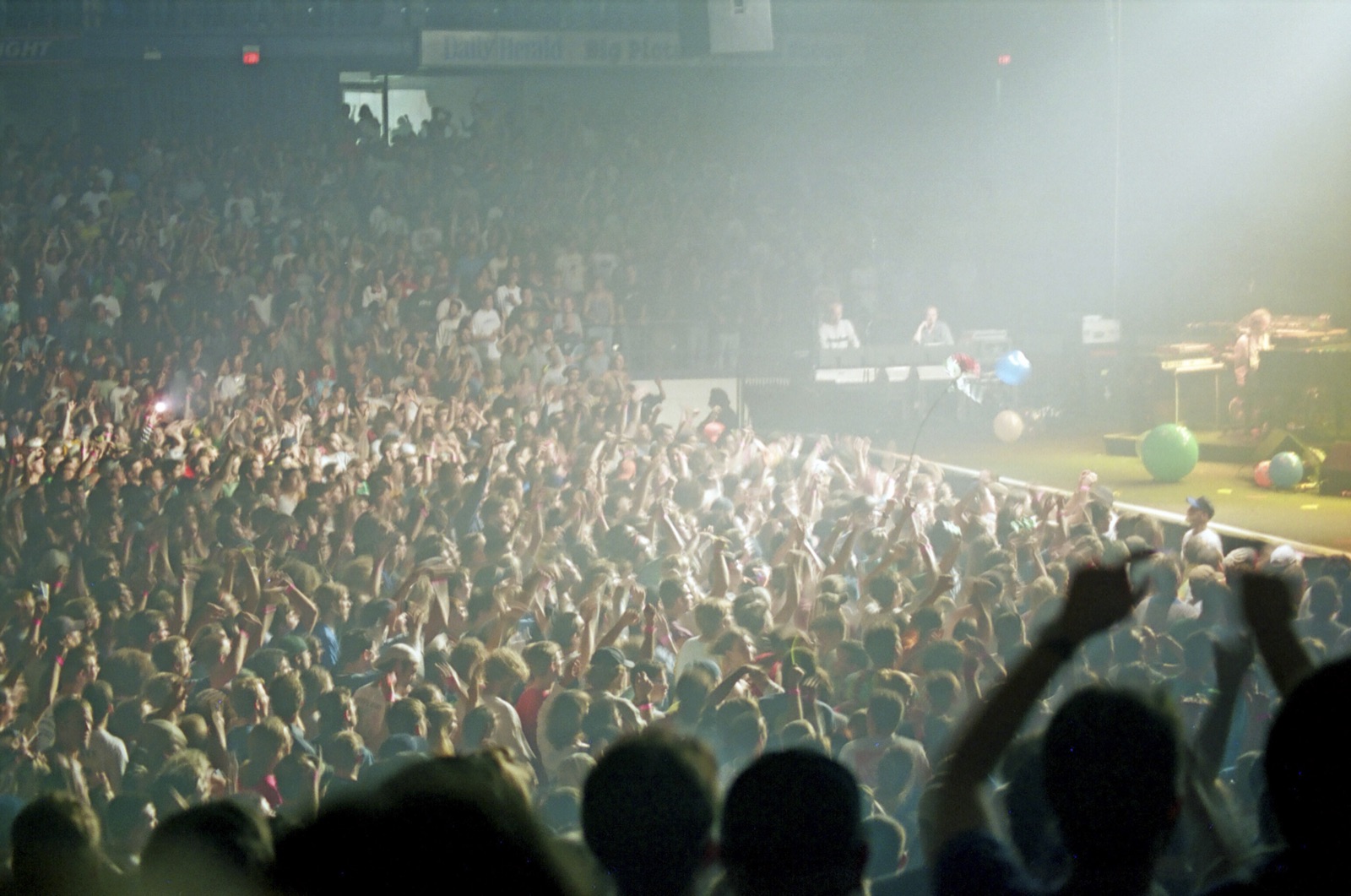 A hazy arena floor full of fans with arms raised toward a stage where beach balls roll by the monitors.